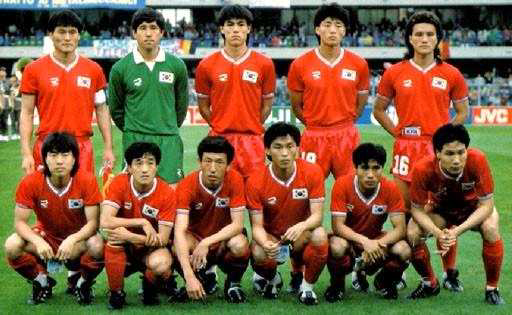 1986년 멕시코 월드컵에 출전한 대한민국 축구 국가대표팀 선수들이 경기 전 단체 사진을 촬영하고 있는 모습. 전원 붉은색 유니폼을 착용하고 있으며, 한 명은 녹색 골키퍼 유니폼을 입고 있다. 상단에는 관중이 보이고, 선수들은 두 줄로 나뉘어 서 있고, 경기장 광고판에는 ‘JVC’ 로고가 보인다.