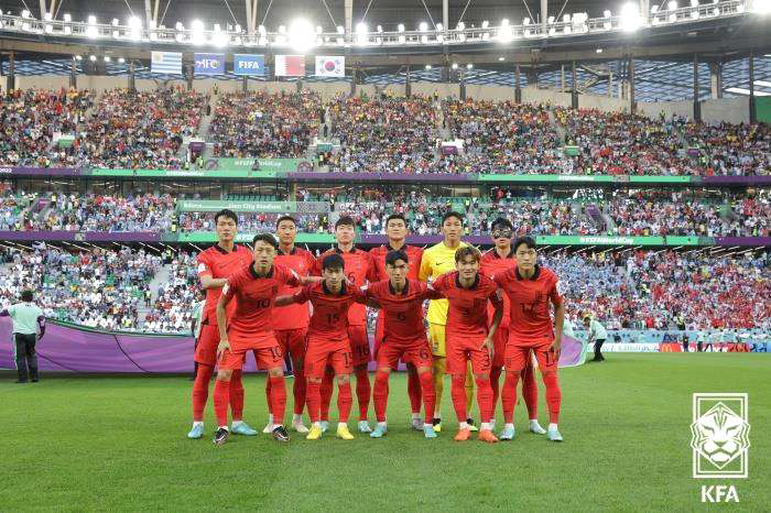 2022 카타르 월드컵 경기 전, 대한민국 축구 국가대표팀이 붉은색 유니폼을 착용하고 단체 사진을 촬영한 모습. 11명의 선발 선수들이 경기장 잔디 위에서 앞줄과 뒷줄로 나뉘어 서 있으며, 경기장 관중석은 각국 팬들로 가득 차 있다. 배경 상단에는 태극기와 FIFA 로고가 보인다. 이미지 우측 하단에는 대한축구협회(KFA) 로고가 삽입되어 있다.