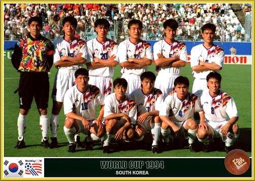 1994년 미국 월드컵에 출전한 대한민국 축구 국가대표팀 선수들이 경기 전 단체 사진을 촬영하고 있는 모습. 선수들은 흰색 유니폼에 붉은색과 파란색의 그래픽 패턴이 들어간 유니폼을 착용하고 있으며, 골키퍼는 다채로운 컬러의 유니폼을 입고 있다. 하단에는 "WORLD CUP 1994 SOUTH KOREA"라는 문구와 함께 FIFA 및 대한민국 국기, 미국 국기 로고가 표시되어 있다. 경기장 배경에는 관중석과 광고판이 보인다.