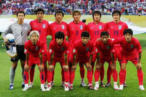 2006년 독일 월드컵에 출전한 대한민국 축구 국가대표팀이 경기 전 단체 기념 촬영을 하고 있다. 붉은색 유니폼을 입은 11명의 선수들이 두 줄로 나뉘어 서 있고, 뒷줄에는 골키퍼를 포함한 선수들이 서 있으며, 앞줄에는 앉은 자세로 포즈를 취하고 있다. 배경에는 경기장을 가득 채운 관중들이 보인다.