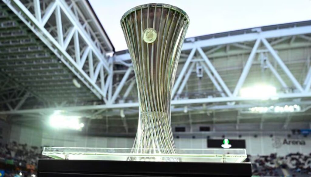 UEFA 유로파 컨퍼런스리그 트로피가 경기장 내 유리 진열대 위에 전시된 모습. 배경은 경기장 관중석과 조명이 비치는 지붕 구조물